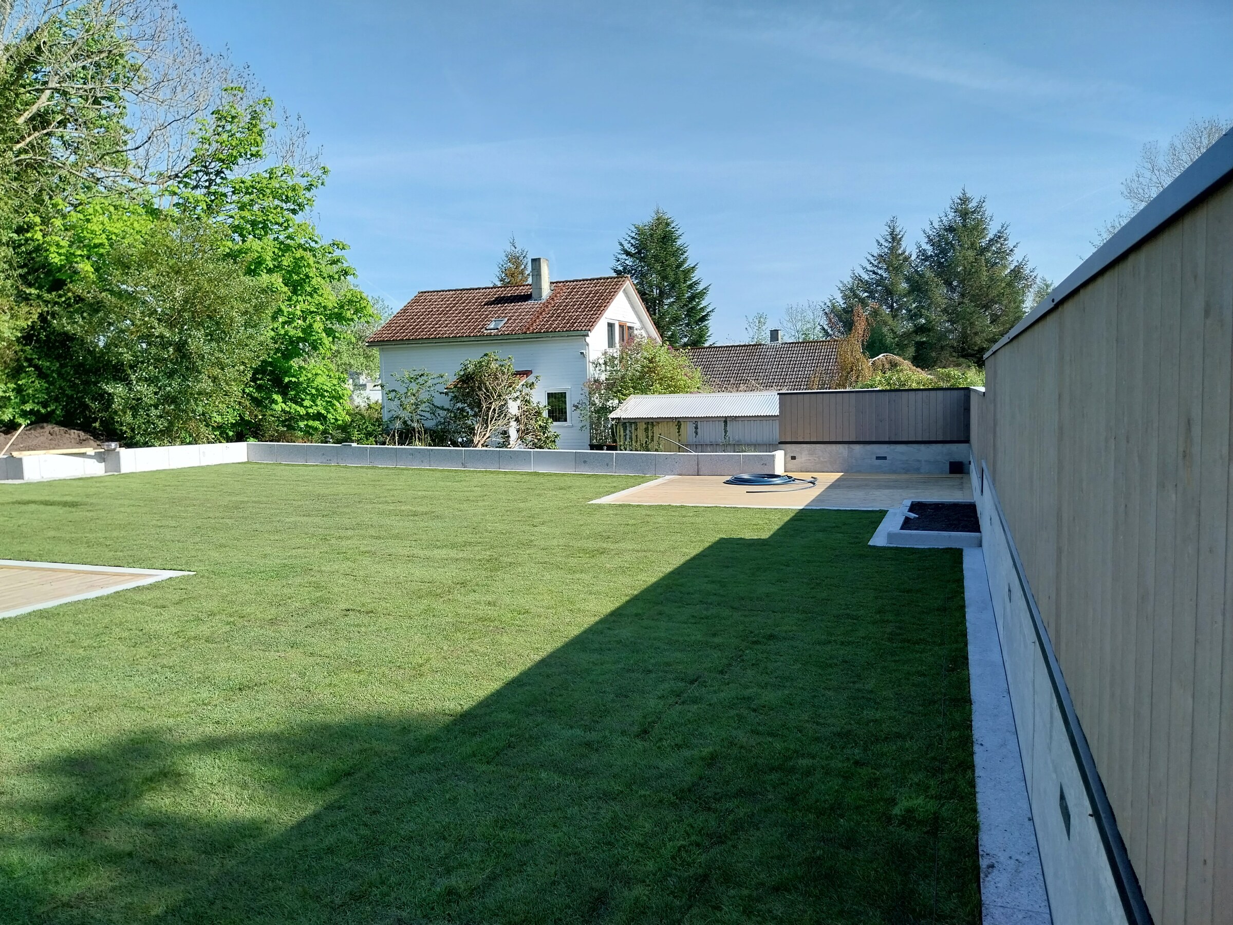 Terrasse i tre og møbler langs husveggen med plenen tett inntil kanten